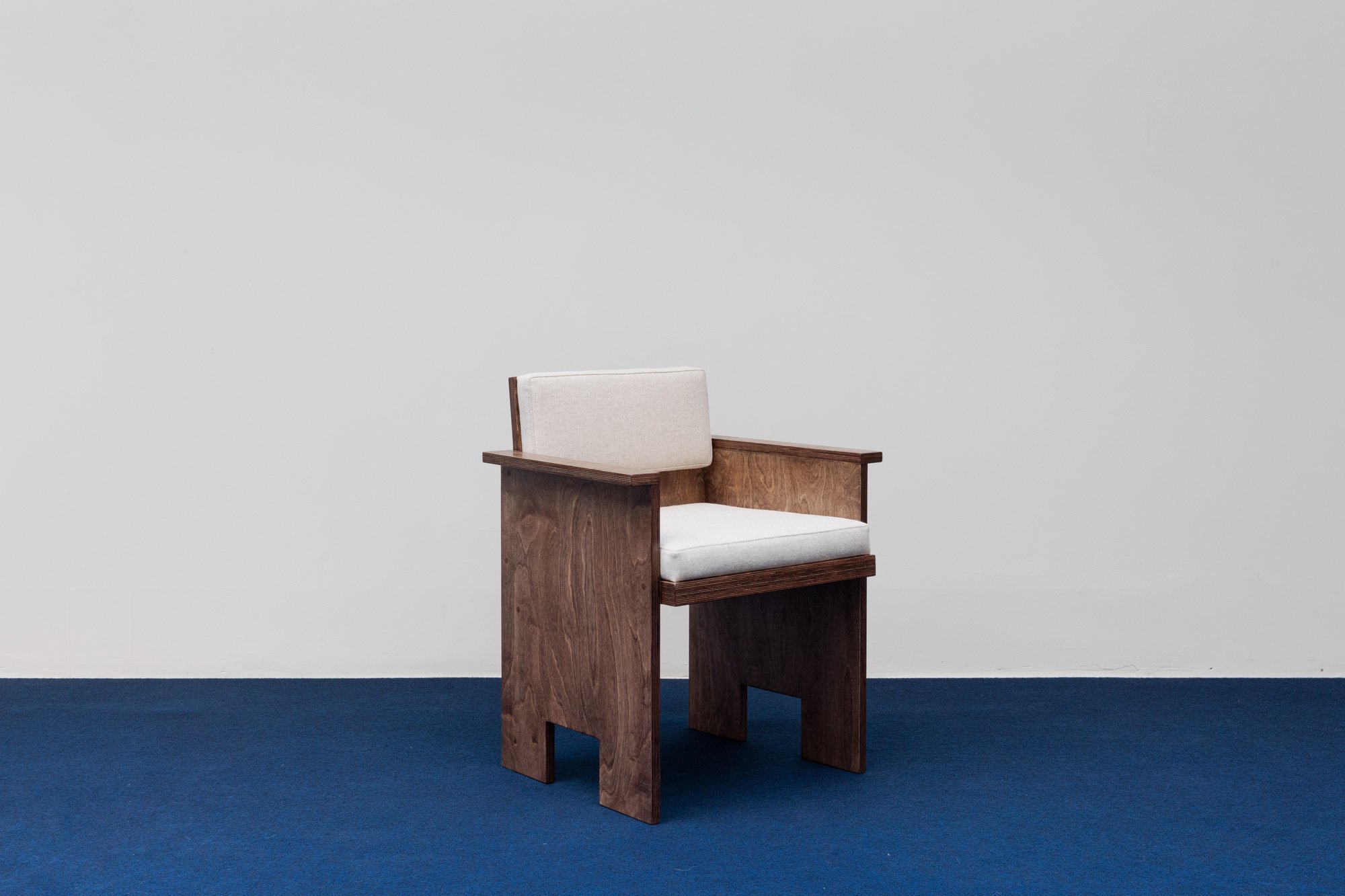 Brace Chair AC by Studio Goons in birch plywood with wool cushions, on a blue carpet against a white wall.
