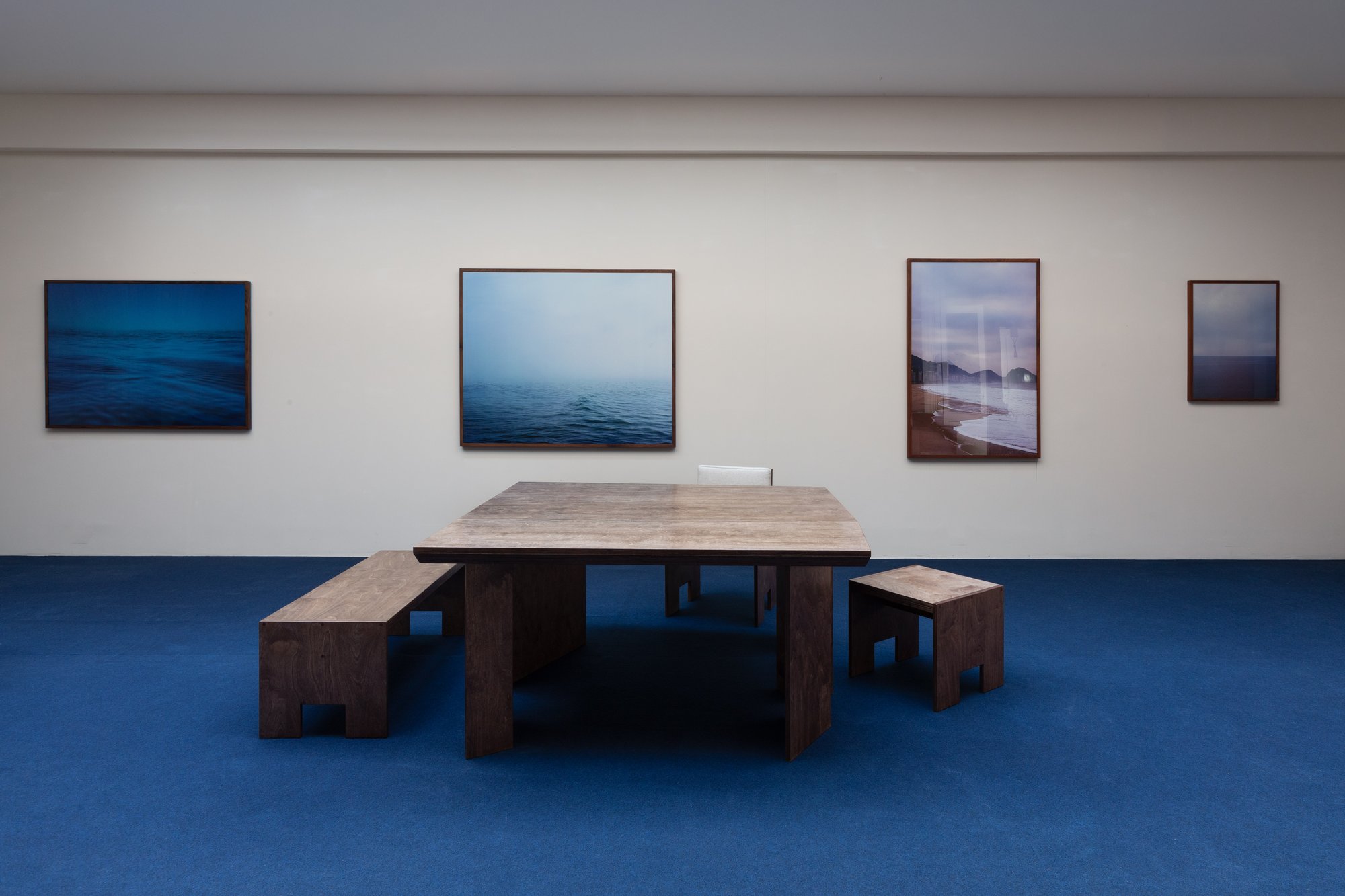 Brace Bench, Brace Stool and Five Points Table by Studio Goons on a blue carpet, with photographs on the gallery wall, Evolving Forms at St Vincents, Antwerp.