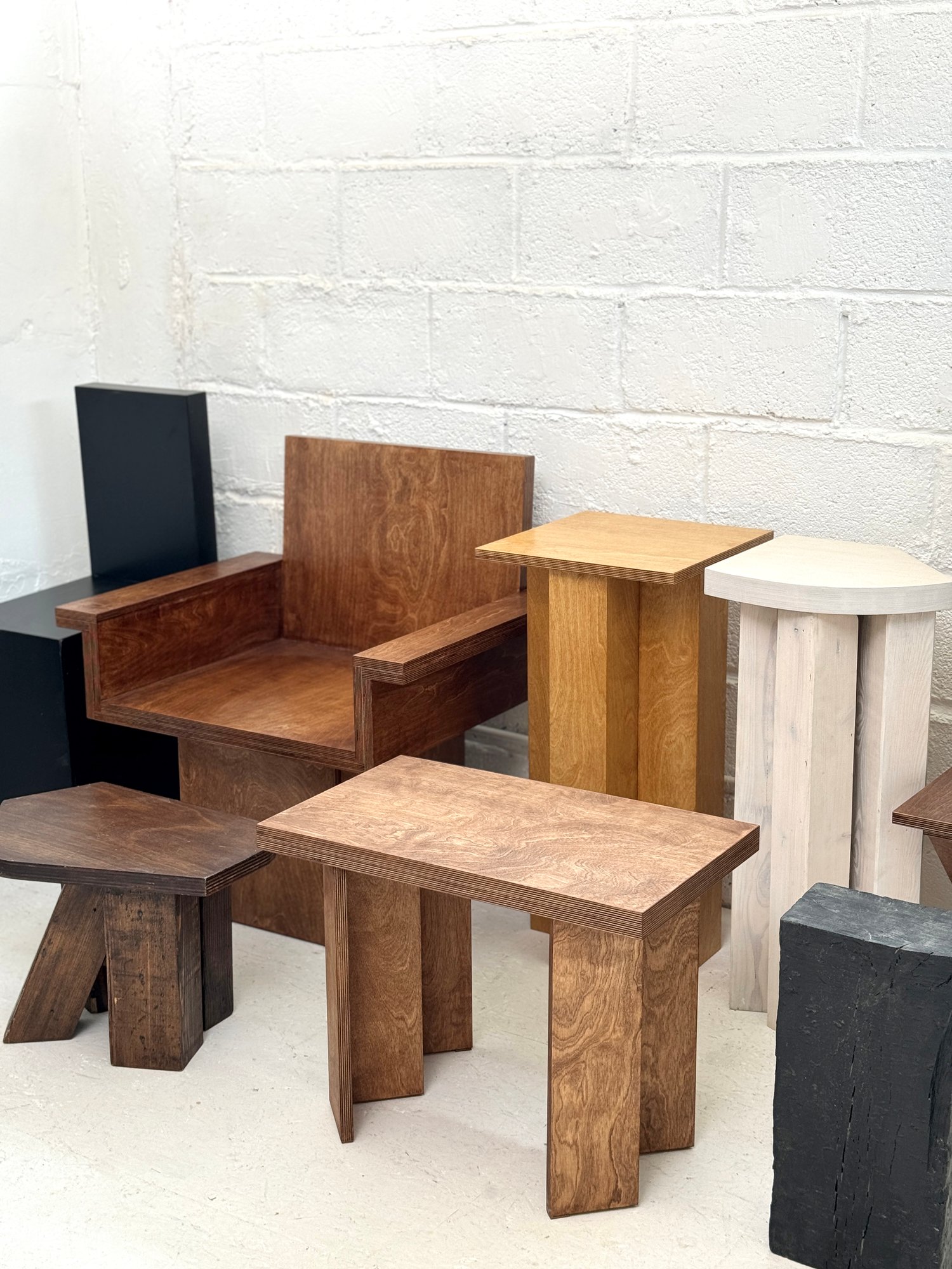 Plywood pieces by Studio Goons in the studio, a black chair, a wood armchair, side tables and stools in several finishes, against a white block wall.