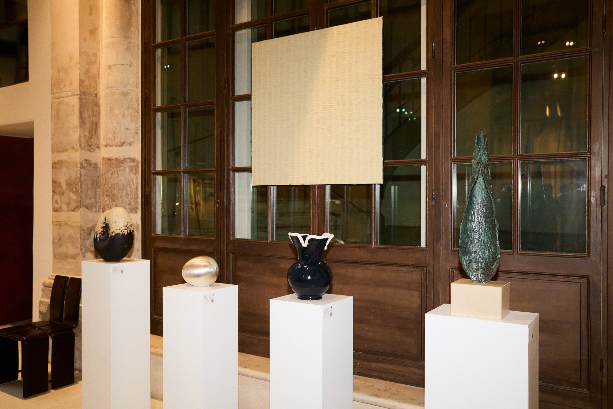 Ceramic and bronze objects on white plinths in front of tall wooden windows at OGATA Paris.